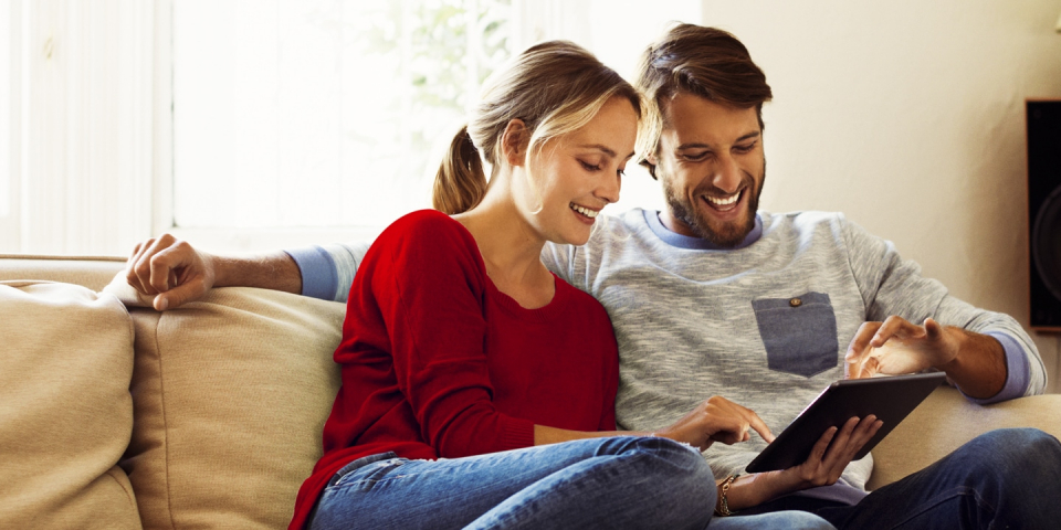 Couple sitting on a sofa at home, looking at a tablet device.