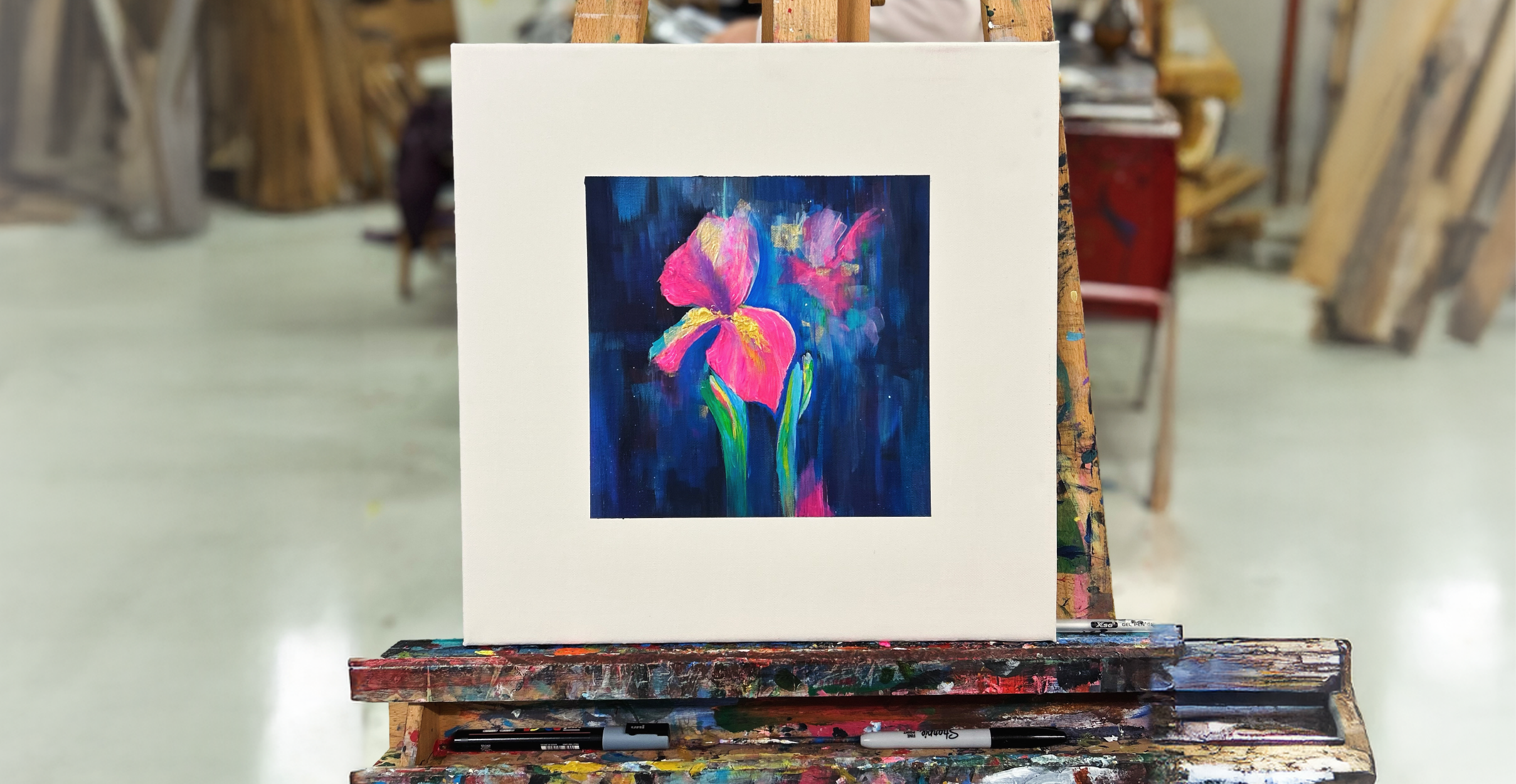 A vibrant painting of a pink and yellow iris with green stems, set against a deep navy and blue textured background. The artwork is displayed on an easel in a studio setting, with brushes, paints, and other tools visible on the easel tray beneath it. This piece, created by David Souch, reflects his passion for exploring colour and form as part of his vision for Overdale Studios, as discussed in the accompanying blog post.